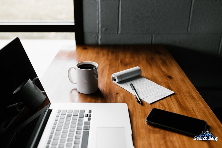 a laptop, notepad, pen, smartphone, and coffee mug sitting on a teakwood table